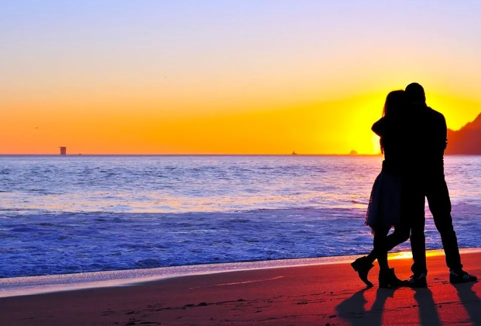 Cận cảnh một cặp đôi đang ôm nhau trìu mến khi nằm trên giường, với ánh sáng ban mai xuyên qua cửa sổ, nhấn mạnh sự ấm áp và gần gũi của lời chúc buổi sáng.