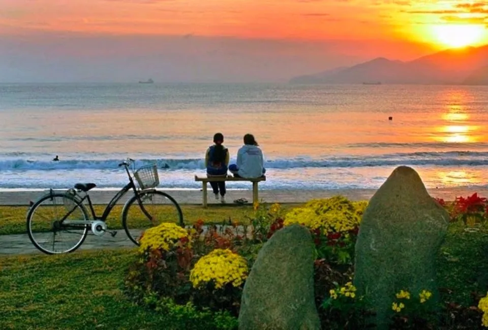 Một người đàn ông đang ôm người yêu của mình từ phía sau, cả hai đang nhìn ra ngoài cửa sổ đón ánh bình minh, thể hiện sự lãng mạn và tình cảm sâu sắc vào buổi sáng.