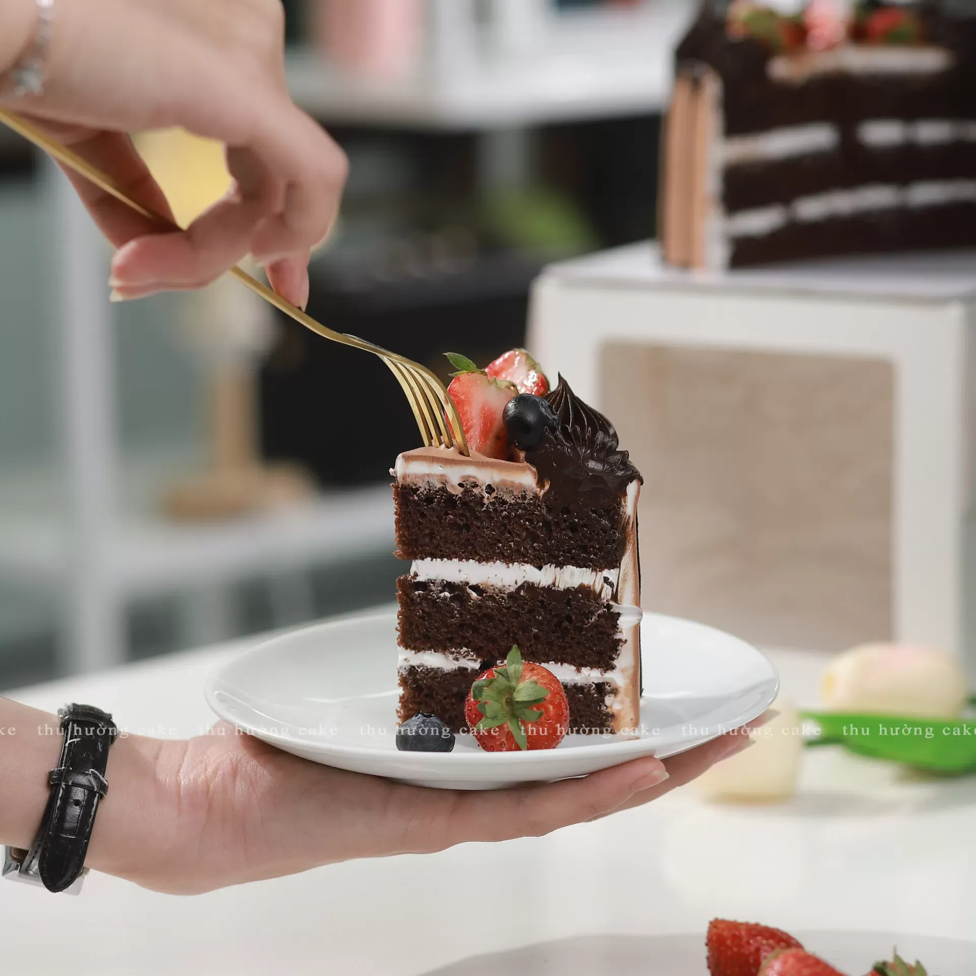 Cốt bánh bông lan vani kích thước 14cm phủ lớp kem socola láng bóng, trang trí dâu tây tươi và ruy băng đỏ đặt trên giá gỗ mộc mạc